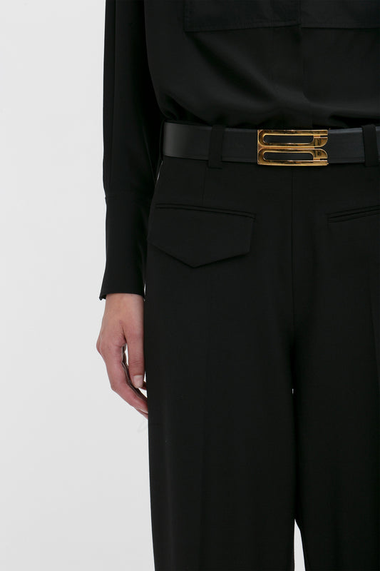 A modern woman is shown from the shoulder to the thigh against a plain white background, donning a black shirt and black pants with a black belt featuring a gold buckle, creating a striking contemporary silhouette with Victoria Beckham's Reverse Front Trouser in Black.