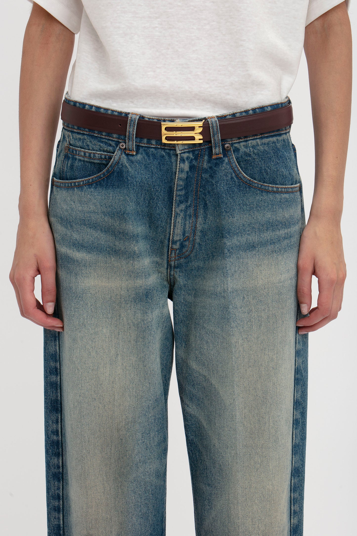 A person wearing blue denim jeans and a white top, with a dark brown Victoria Beckham Frame Belt In Burgundy Leather featuring gold hardware. The view is cropped to show the waist and upper legs.