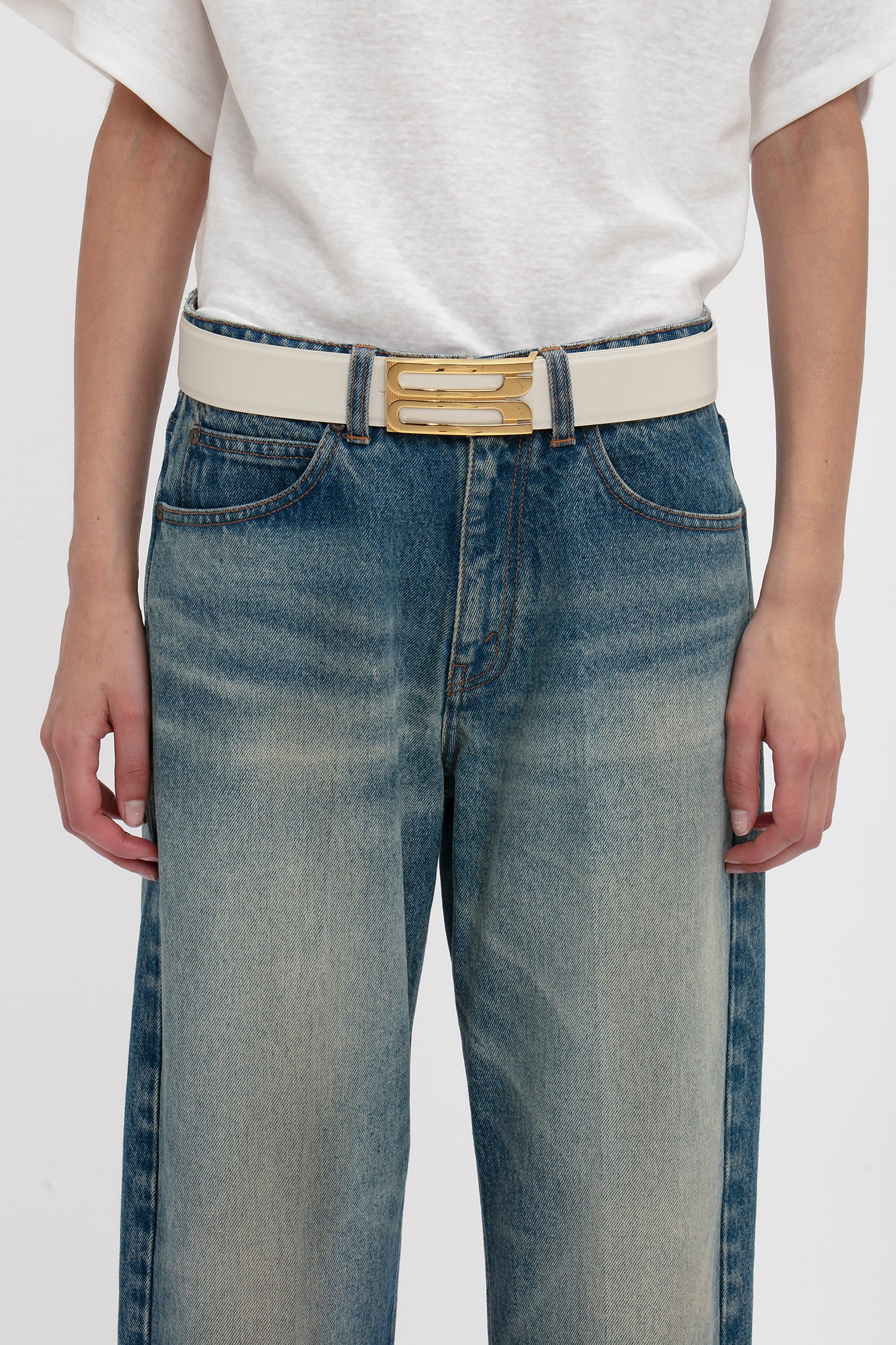 A person is wearing a white t-shirt tucked into faded blue jeans, secured with a Victoria Beckham Jumbo Frame Belt In Latte Leather featuring gold hardware.