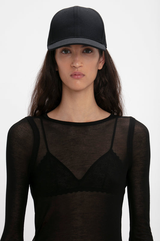 Woman wearing a sheer black top and a Victoria Beckham Exclusive Logo Cap In Black, standing against a white background, gazing directly at the camera.
