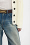 Close-up of a person wearing a Relaxed Fit Cardigan In Natural by Victoria Beckham, a ribbed white shirt, and distressed blue jeans with a brown belt that has a unique, layered gold buckle.