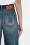 A person wearing blue denim Alina High Waisted Jean In Indigrey Wash by Victoria Beckham with seventies-style pockets and a brown leather patch on the waistband is shown from the back. The right arm hangs by their side.