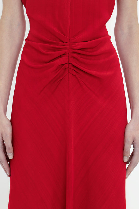 Close-up of a person wearing a red Exclusive Gathered Waist Midi Dress In Carmine by Victoria Beckham, with ruched detailing around the waist, arms resting by sides.