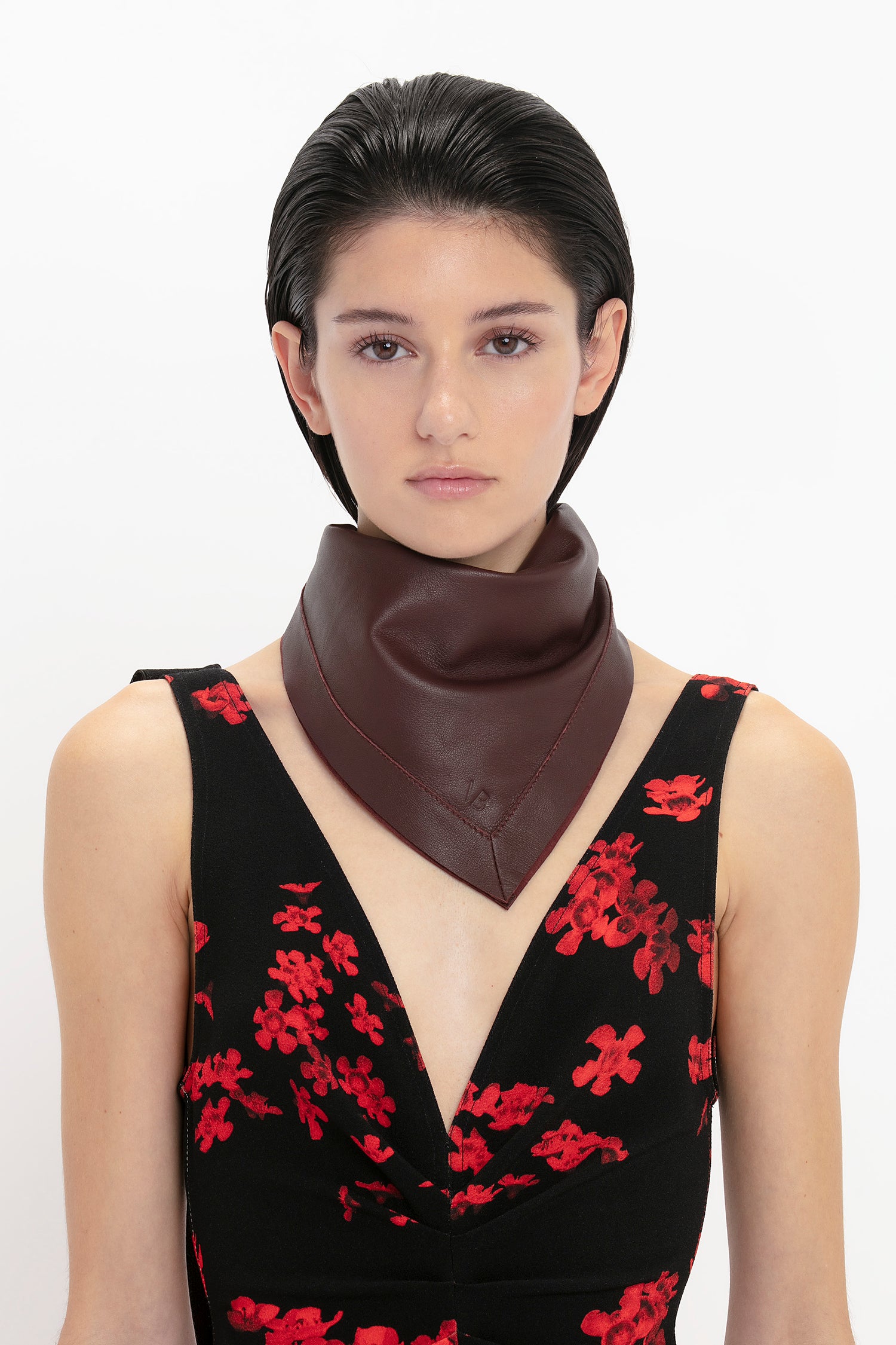 A person with short dark hair wears a black dress with red floral patterns and a Leather Scarf In Bordeaux by Victoria Beckham. They stand against a plain white background.