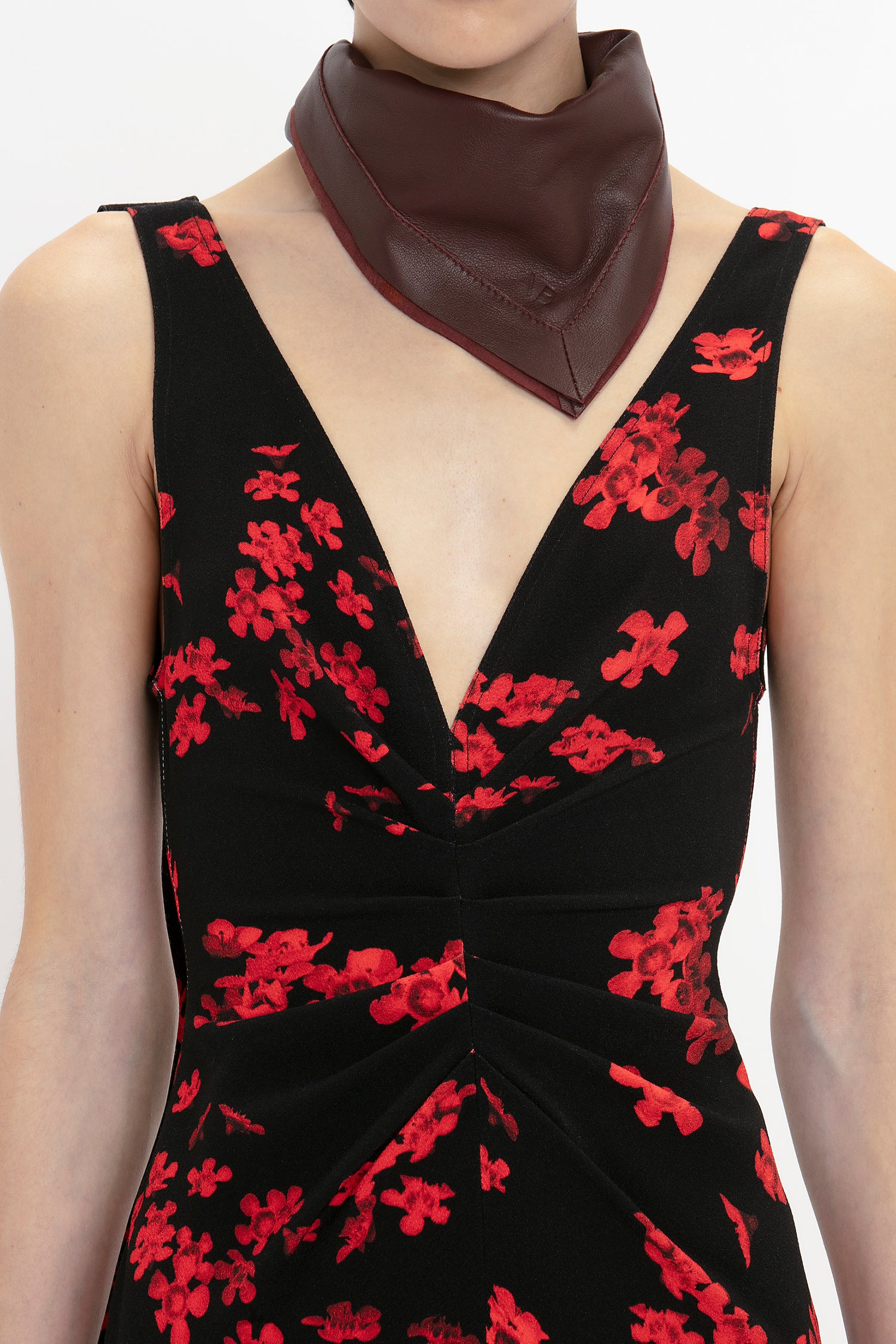 A person wears a black dress with red floral patterns and a Victoria Beckham Leather Scarf In Bordeaux. The dress, featuring a deep V-neckline, perfectly balances between feminine separates and masculine tailoring.