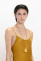 A person with short dark hair wearing a Victoria Beckham Low Back Cami Floor-Length Dress in Harvest Gold and a long gold necklace with a rectangular pendant stands against a plain white background.