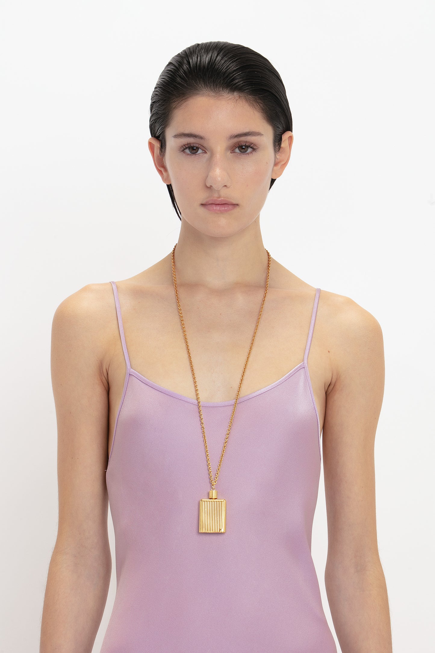 Person with short dark hair wearing a Victoria Beckham Low Back Cami Floor-Length Dress In Rosa and a long gold necklace with a square pendant, creating a sensuous silhouette against the plain white background.