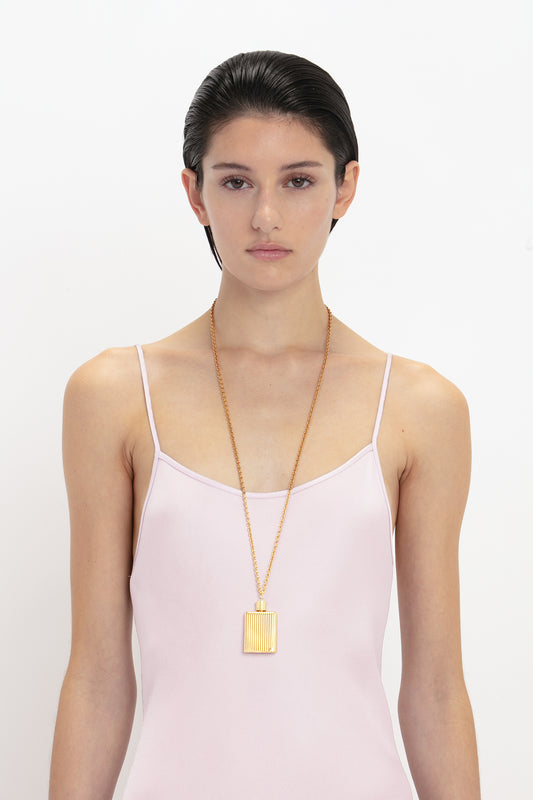 A woman with slicked-back hair wearing a Victoria Beckham Low Back Cami Floor-Length Dress In Rosa and a rectangular gold pendant necklace, standing against a white background.