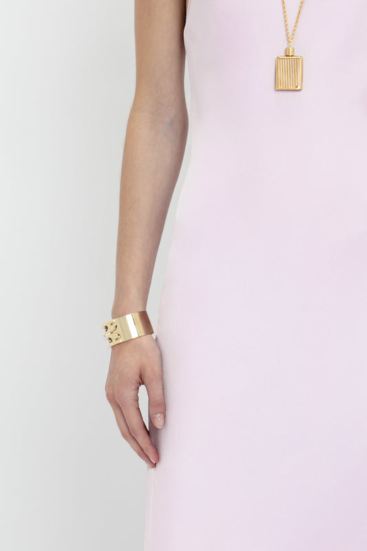 A person wearing a Victoria Beckham Low Back Cami Floor-Length Dress In Rosa with a gold cuff bracelet and a rectangular pendant necklace against a gray background.