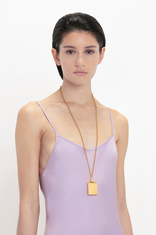 A person in a Victoria Beckham Low Back Cami Floor-Length Dress In Petunia and a long gold necklace with a rectangular pendant stands against a white background, evoking 1990s style.
