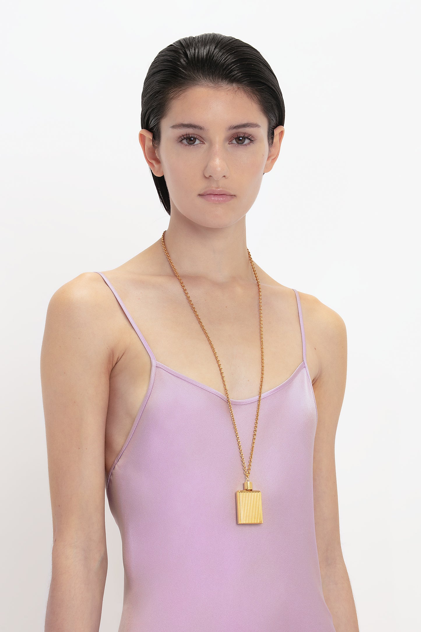 A person with short dark hair is wearing a Low Back Cami Floor-Length Dress In Rosa by Victoria Beckham and a long gold necklace with a rectangular pendant, standing against a plain white background.