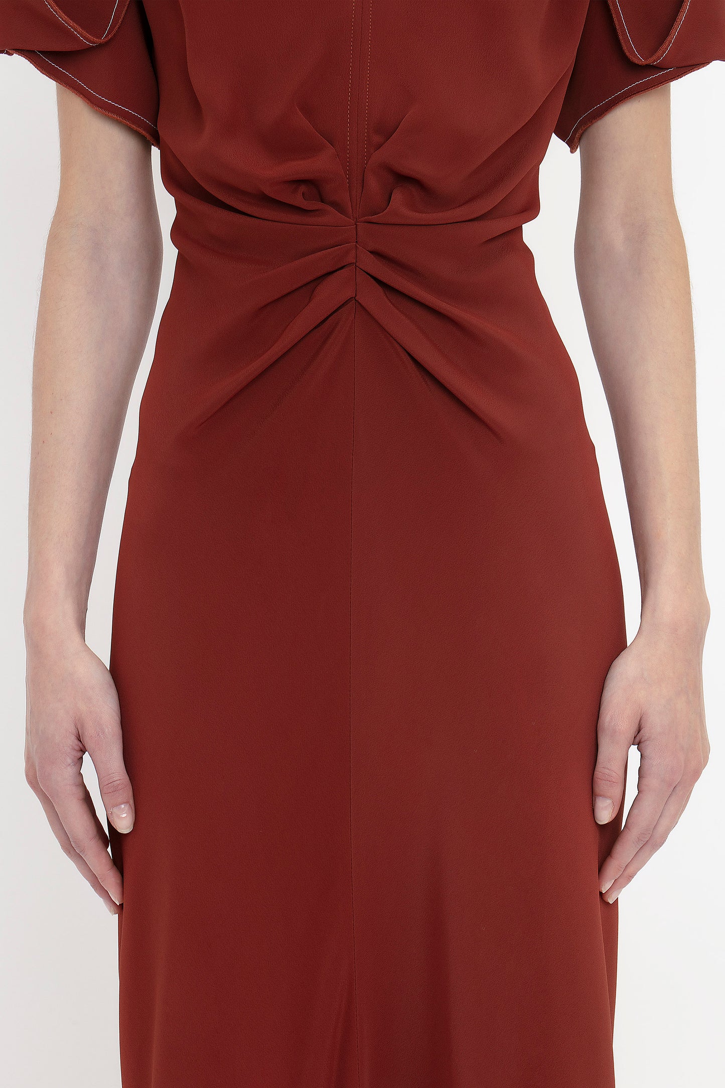 A person is wearing a rust-colored Gathered V-Neck Midi Dress In Russet by Victoria Beckham, with waist-defining pleat detail and slightly flared short sleeves, standing against a white background. Their hands are relaxed by their sides.