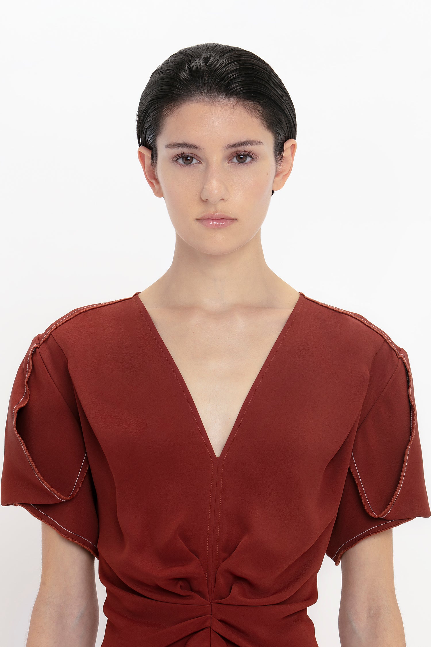 Person with short dark hair wearing a Victoria Beckham Gathered V-Neck Midi Dress in Russet, standing against a plain white background.