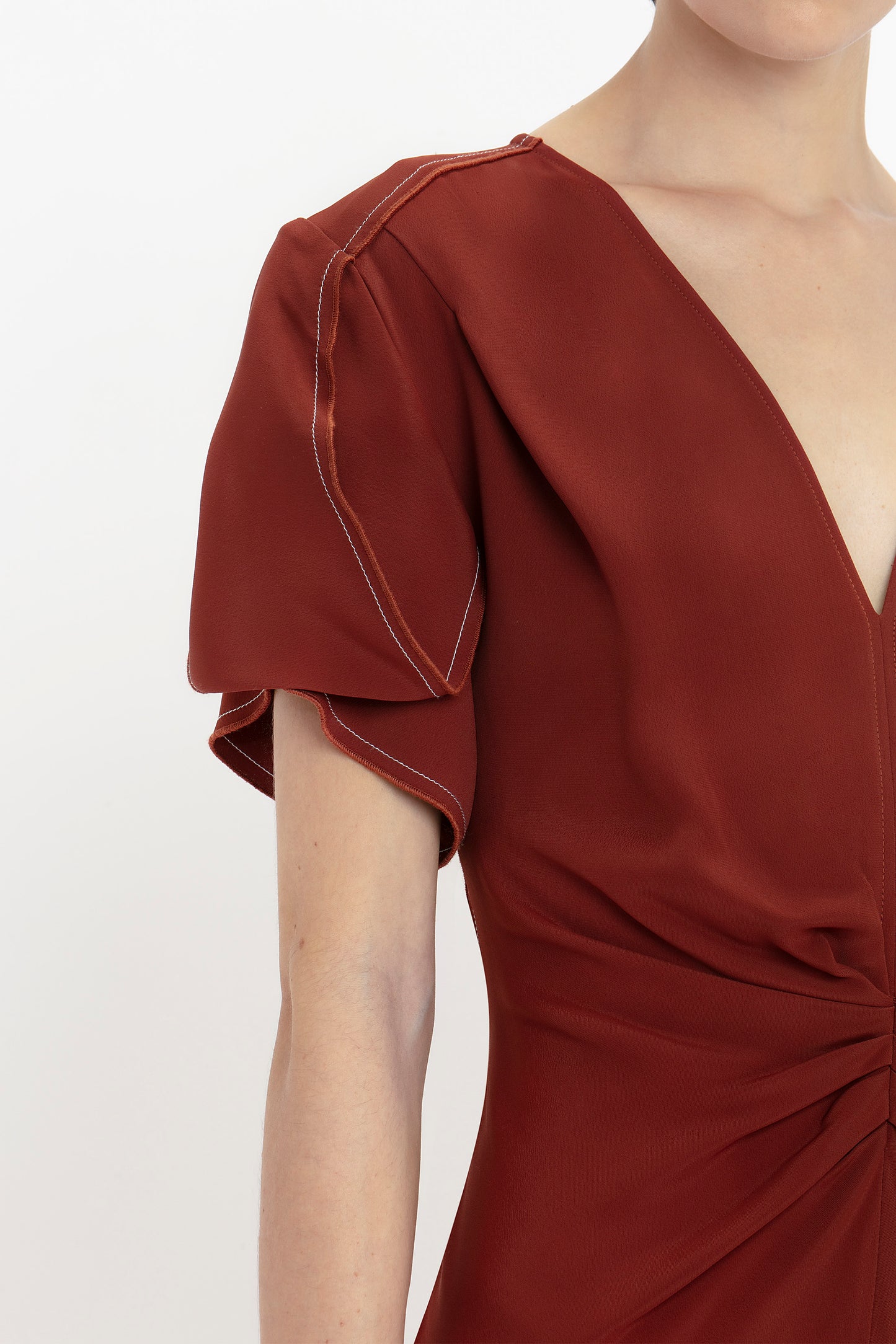 Close-up of a person wearing the Gathered V-Neck Midi Dress In Russet by Victoria Beckham, a waist-defining pleat detail dress with a deep V-neckline and short puff sleeves, featuring white stitching details.