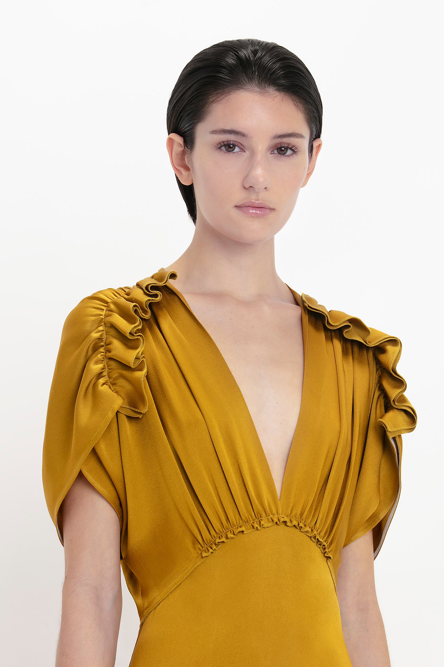 A person with short dark hair wears a V-Neck Ruffle Midi Dress In Harvest Gold by Victoria Beckham, featuring shoulder frills, voluminous ruffled sleeves, and a deep V-neckline. The background is plain white.