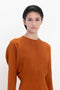 A person with short dark hair is wearing an orange Dolman Midi Dress In Russet by Victoria Beckham, crafted from fluid cady fabric, looking at the camera with a neutral expression.