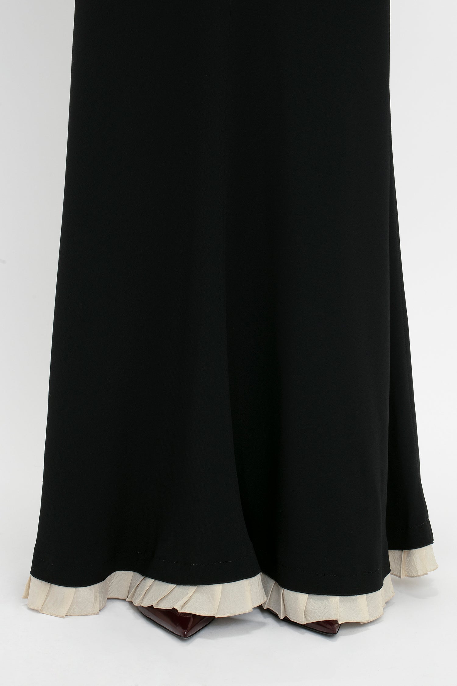 Close-up of the hem of a Victoria Beckham V-Neck Gathered Waist Floor-Length Gown In Black with a white frilled edge. The dress is floor-length and partially obstructs a pair of dark-colored shoes.