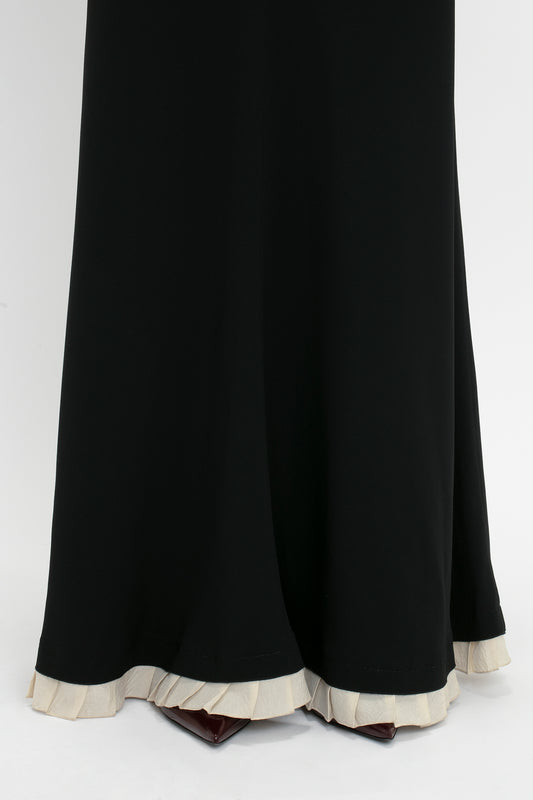 Close-up of the hem of a Victoria Beckham V-Neck Gathered Waist Floor-Length Gown In Black with a white frilled edge. The dress is floor-length and partially obstructs a pair of dark-colored shoes.
