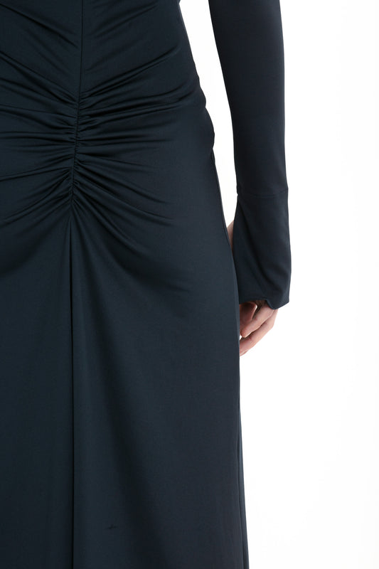 Close-up of a person wearing an evening gown with understated glamour—a long-sleeved, ruched black dress. The image captures the back view from the waist downwards, their right hand resting beside the Victoria Beckham Ruched Detail Floor-Length Gown In Midnight.