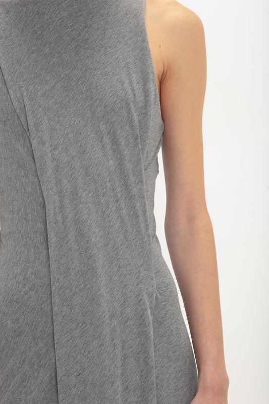 Close-up of a person’s torso and arm wearing a sleeveless, asymmetrical, grey Frame Detailed Maxi Dress In Titanium by Victoria Beckham with an elongated silhouette against a white background. The person's face and lower body are not visible.