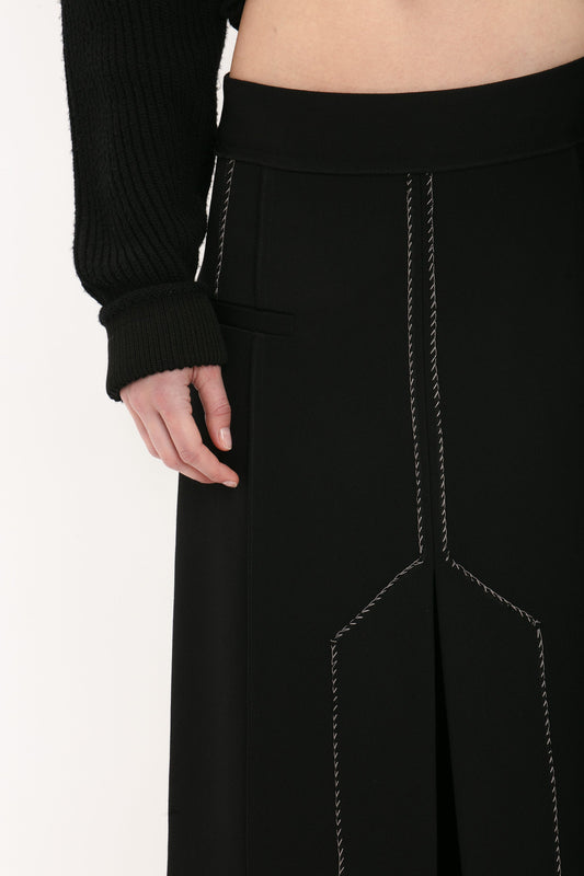 Person wearing a black long-sleeve top and black pants with white stitching detail, showcasing an elongated silhouette, with their hand resting by the side is actually sporting the Deconstructed Floor-Length Skirt In Black by Victoria Beckham.