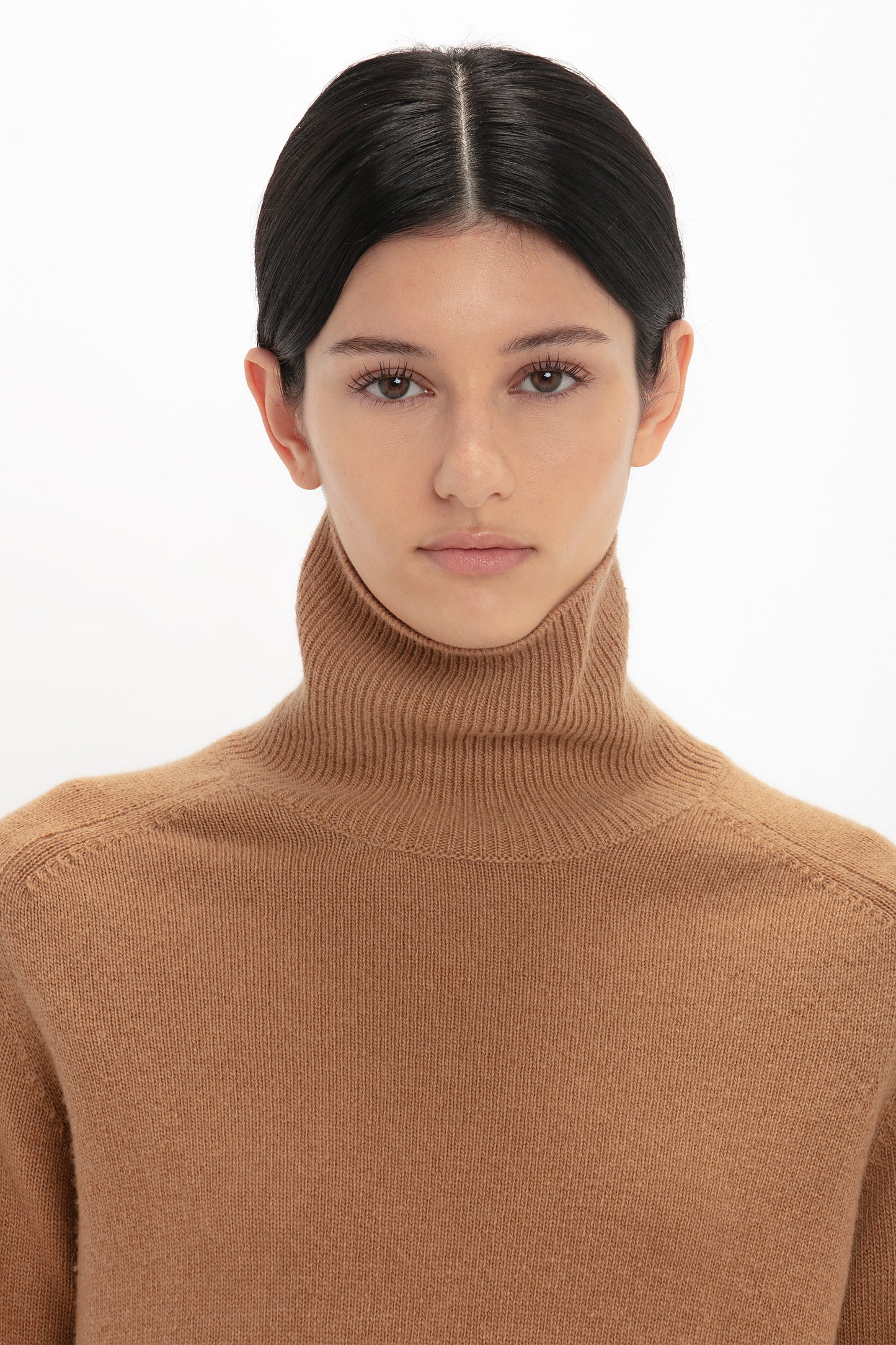 A person with dark hair is wearing a Victoria Beckham Polo Neck Jumper In Tobacco and looking directly at the camera. The background is white.