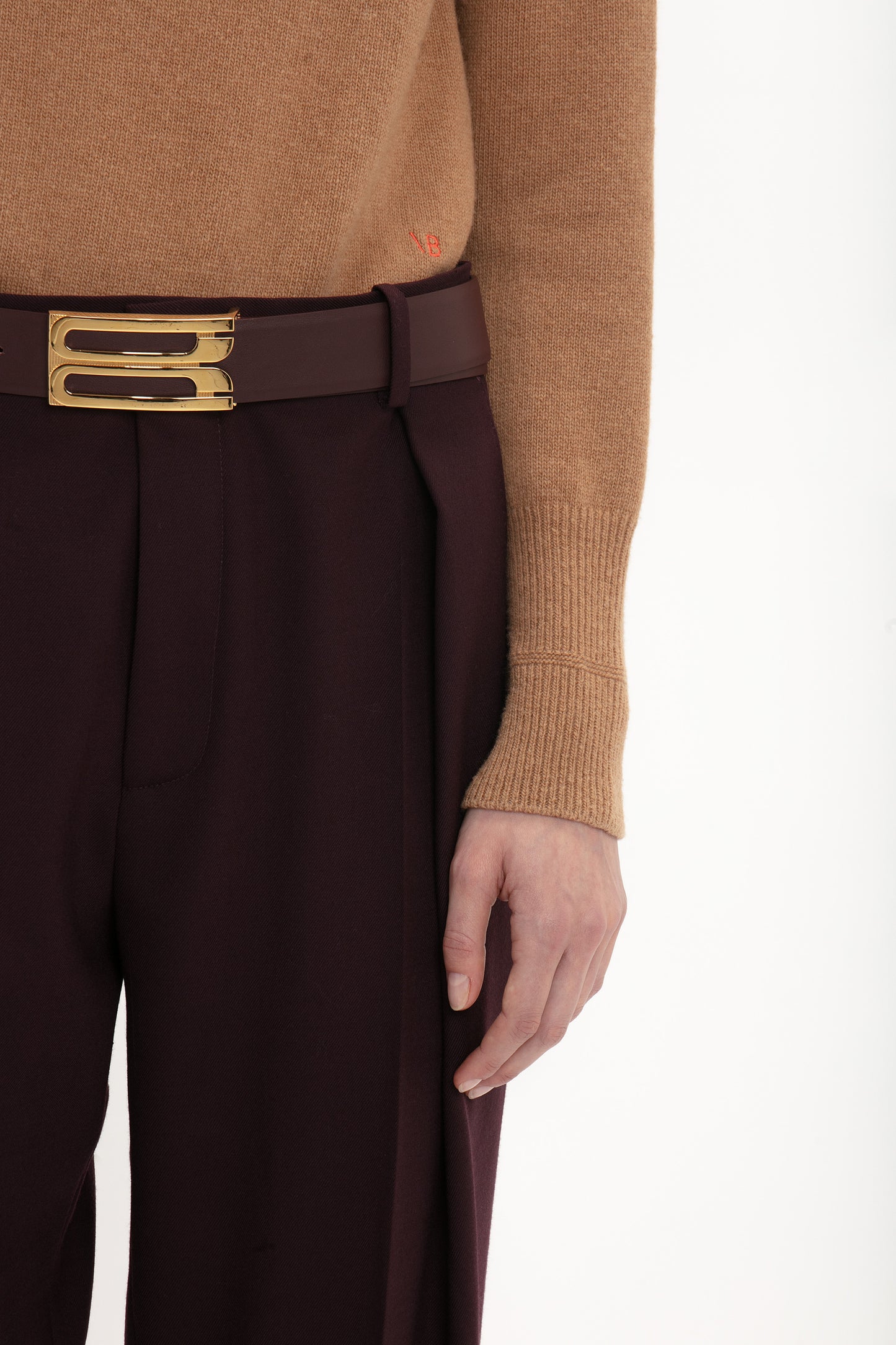 A person wearing luxury knitwear—a Victoria Beckham Polo Neck Jumper In Tobacco—and maroon pants with a gold belt buckle, showing their right arm and hand.