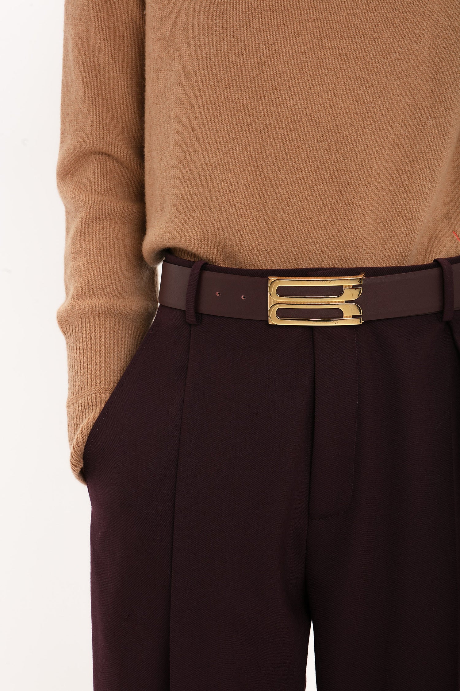 Person wearing a brown sweater tucked into high-waisted Victoria Beckham Asymmetric Chino Trouser In Deep Mahogany with a matching maroon belt featuring a gold buckle, showcasing a narrow leg silhouette.