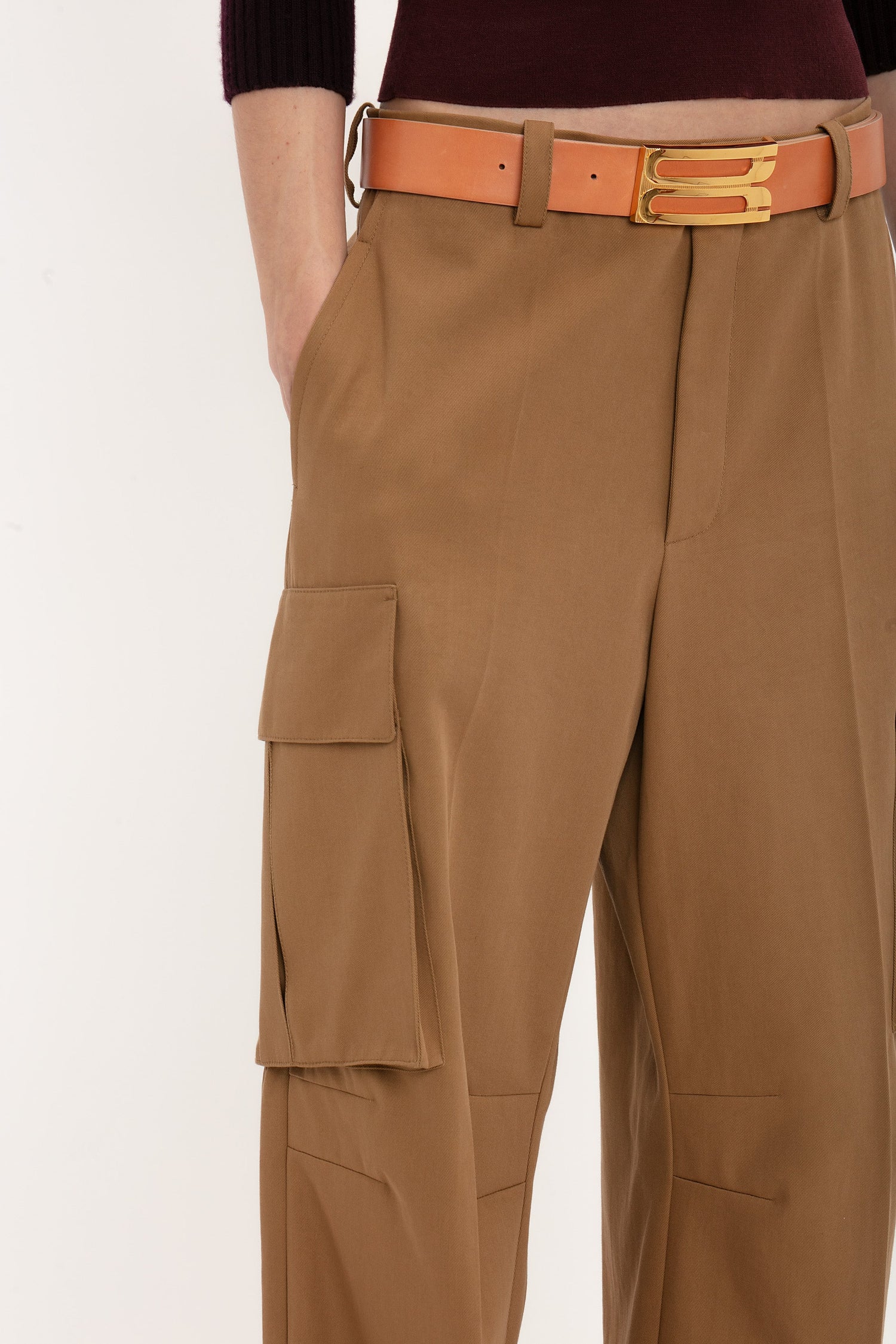 Close-up of a person wearing the Victoria Beckham Relaxed Cargo Trouser In Tobacco with large pockets and a tan belt featuring a gold buckle. The person's hand rests in the pocket, accentuating the military-inspired style. The top part of a dark-colored sweater is partially visible, adding to the relaxed silhouette.