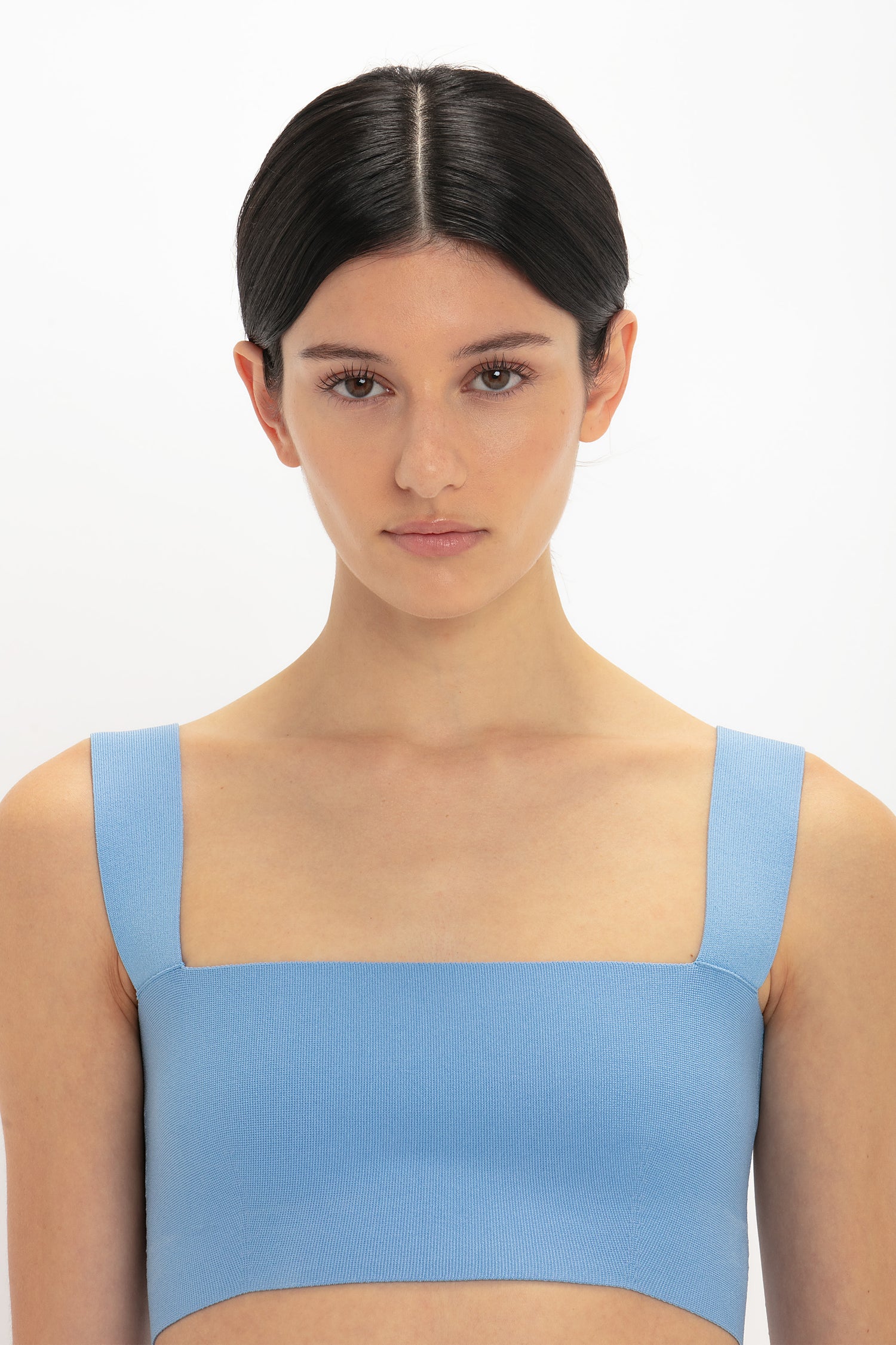 A person with dark hair styled in a middle part is wearing an ultra form-fitting, light blue Strap Bandeau Top In Marina by Victoria Beckham, looking directly at the camera against a plain white background.