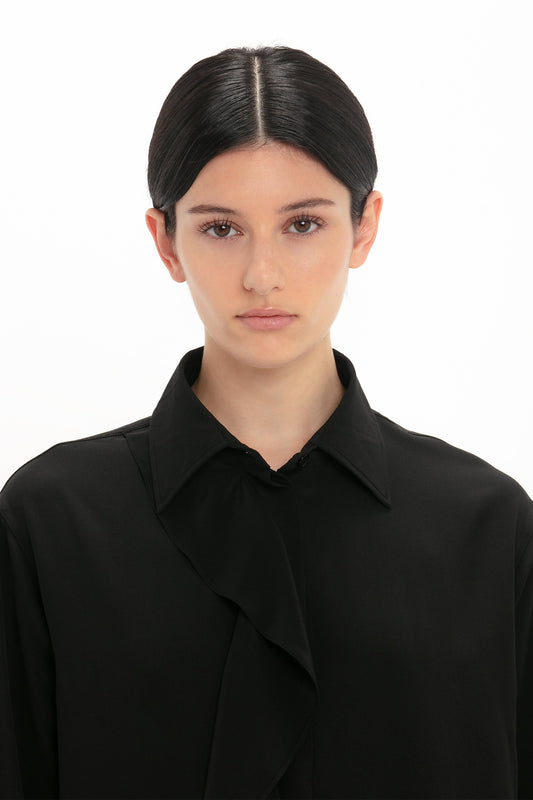 A person with dark hair is wearing a Victoria Beckham Asymmetric Ruffle Blouse In Black. They are facing the camera with a neutral expression against a plain white background.
