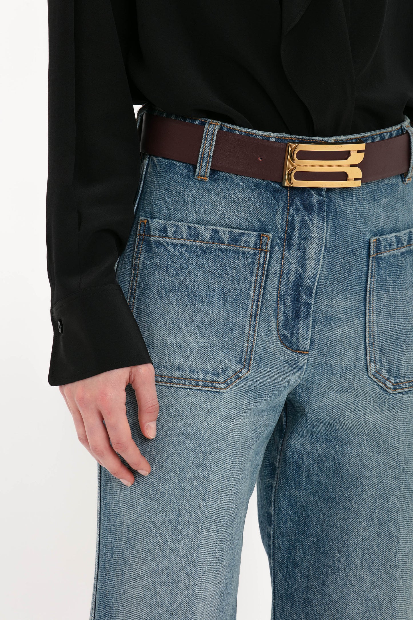 A person wearing a black long-sleeve shirt, Victoria Beckham Alina High Waisted Jean In Worn Blue Wash, and a brown belt with a metallic buckle.