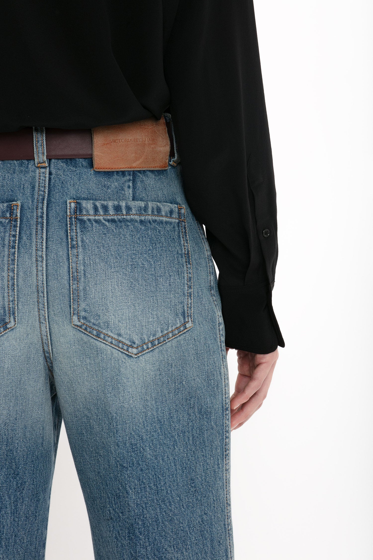 Back view of a person wearing a black shirt tucked into blue denim Victoria Beckham Alina High Waisted Jean In Worn Blue Wash with seventies-inspired patch pockets and a distressed Worn Blue Wash, featuring a brown leather patch on the waistband.