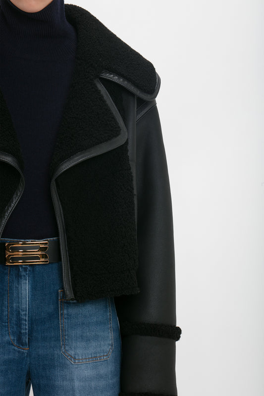 Person wearing a Victoria Beckham Shearling Jacket In Black, dark turtleneck, and blue jeans with a gold-buckled belt. Only the upper torso is visible.