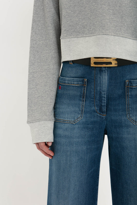 Close-up of a person wearing blue jeans and a Victoria Beckham Cropped Sweatshirt In Grey Marl, hand resting on hip, with a partial view of a black belt.