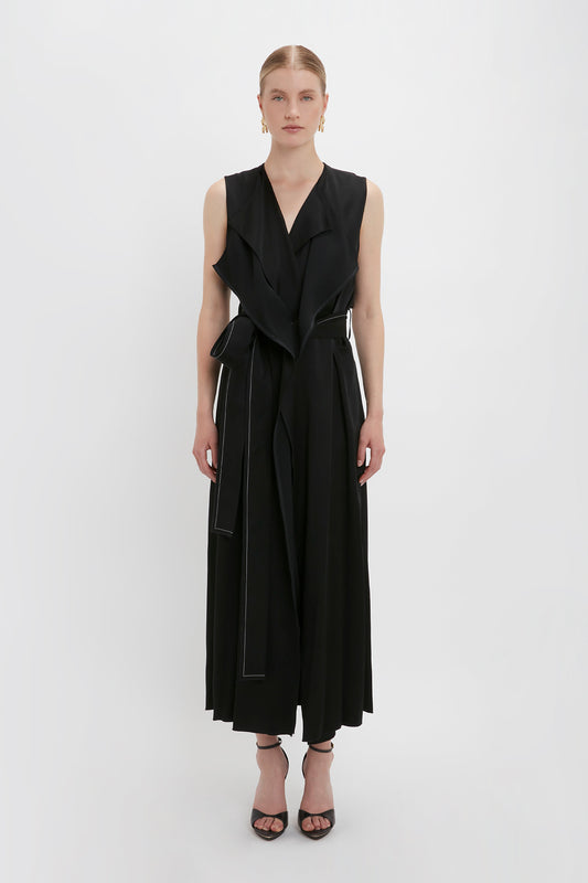 A woman stands against a white background wearing a sleeveless, black wrap-front Trench Dress In Black by Victoria Beckham with wide legs, a sash-style belt, oversized front flap pockets, and black high-heeled sandals.