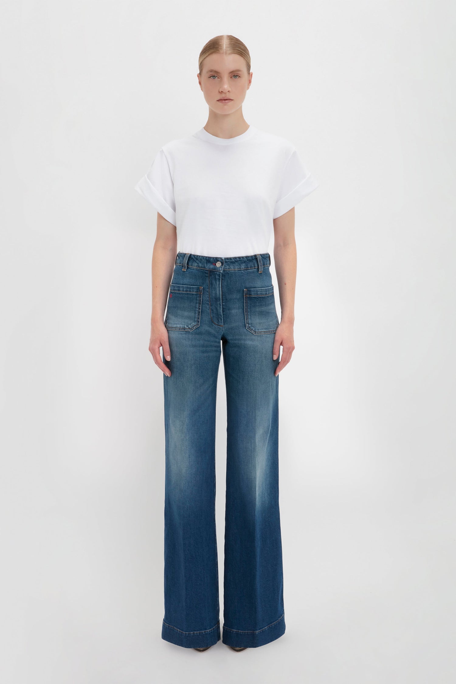 A woman standing against a white background wearing an oversized Asymmetric Relaxed Fit T-Shirt in White and blue wide-leg jeans by Victoria Beckham.