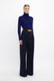 A person stands against a plain background wearing a blue turtleneck sweater, Victoria Beckham Wide Leg Trouser In Midnight with an asymmetric zip placket, and a belt with a gold buckle.