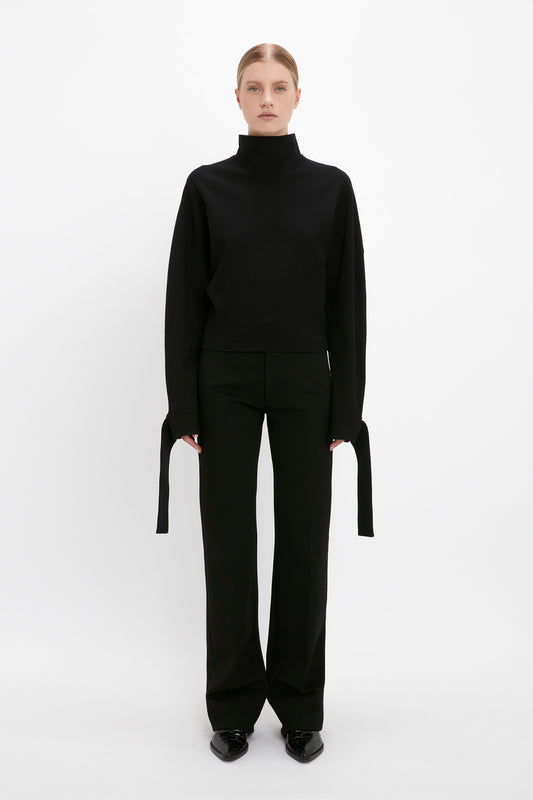 A person stands against a plain white background, effortlessly showcasing off-duty dressing in a Victoria Beckham Tie Sleeve Ponti Top In Black and black pants, with their hands hanging by their sides.