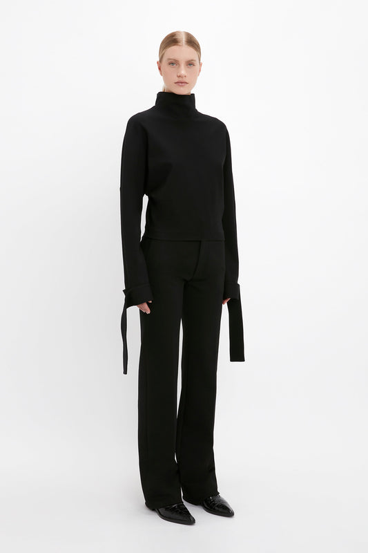 Person showcasing off-duty dressing with the Victoria Beckham Tie Sleeve Ponti Top In Black featuring long sleeves and black straight-leg pants, standing against a plain white background.