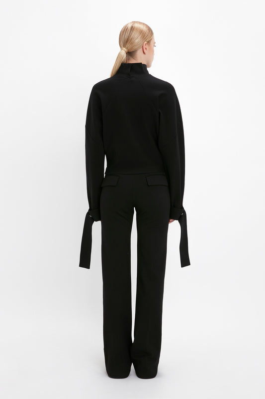 A person with blonde hair is standing with their back to the camera, wearing a black Tie Sleeve Ponti Top In Black by Victoria Beckham and long pants, perfect for off-duty dressing.