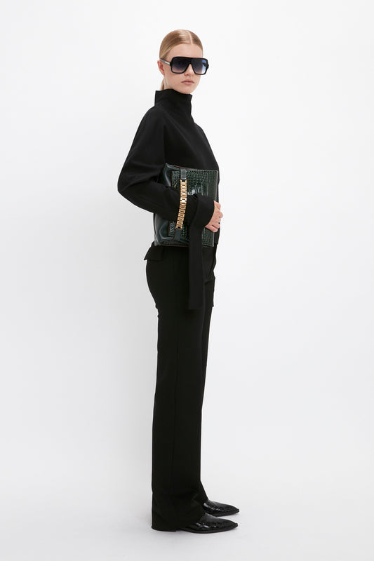 Woman in a Tie Sleeve Ponti Top In Black by Victoria Beckham, large sunglasses, and black shoes stands against a white background, holding a green crocodile-embossed bag with a gold chain strap—epitomizing effortless off-duty dressing.