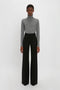 Person wearing a Victoria Beckham Polo Neck Jumper In Grey Melange and black wide-leg trousers stands against a plain white background.