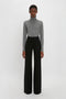 Person stands against a plain white background wearing a gray turtleneck sweater and black Alina Tailored Trouser In Black by Victoria Beckham with super-high waist and flared legs.