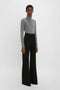 A person in a grey turtleneck sweater and Victoria Beckham's Alina Tailored Trouser In Black with a super-high waist and flared leg stands against a plain white background.