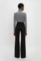 A person facing away from the camera wears a grey turtleneck sweater and black Alina Tailored Trouser In Black by Victoria Beckham with a super-high waist against a plain white background.