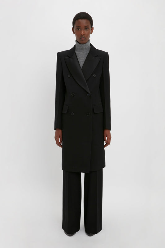 An individual stands against a plain white background wearing a slim fit, black Double Breasted Tuxedo Coat in Black by Victoria Beckham over a gray turtleneck and black trousers.