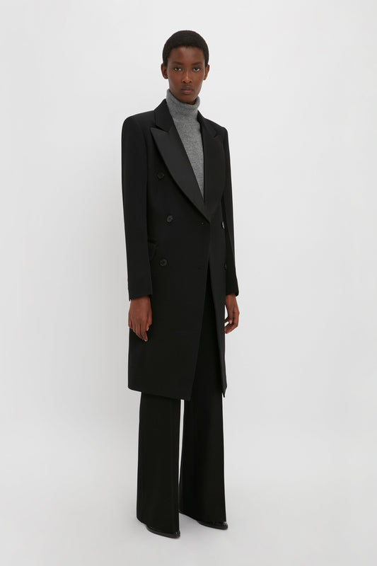 A person stands against a plain background wearing a Victoria Beckham Double Breasted Tuxedo Coat in Black, gray turtleneck, and black wide-leg pants.