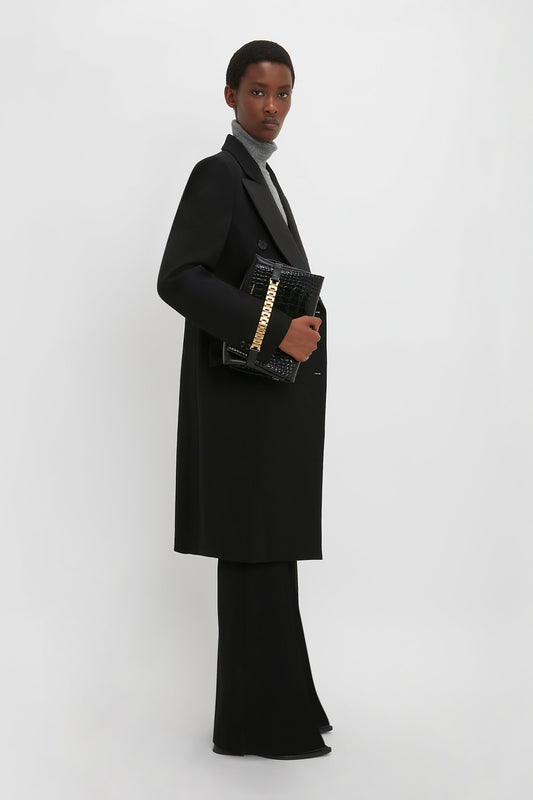 A person stands against a white background, wearing a black wool gabardine Double Breasted Tuxedo Coat in Black by Victoria Beckham, a grey top, and slim fit black pants. They hold a black textured clutch bag with a gold chain detail.