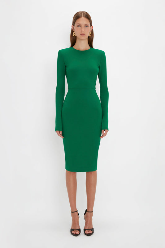 A person stands against a plain white background wearing the Victoria Beckham Long Sleeve T-Shirt Fitted Dress in Emerald and black heels.